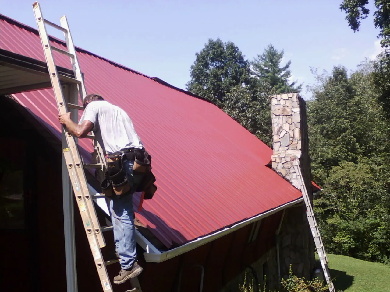 Roofer on a ladder beside a red metal roof and stone chimney — Soffit & Fascia Repair by Roofing Brothers in Middlesboro